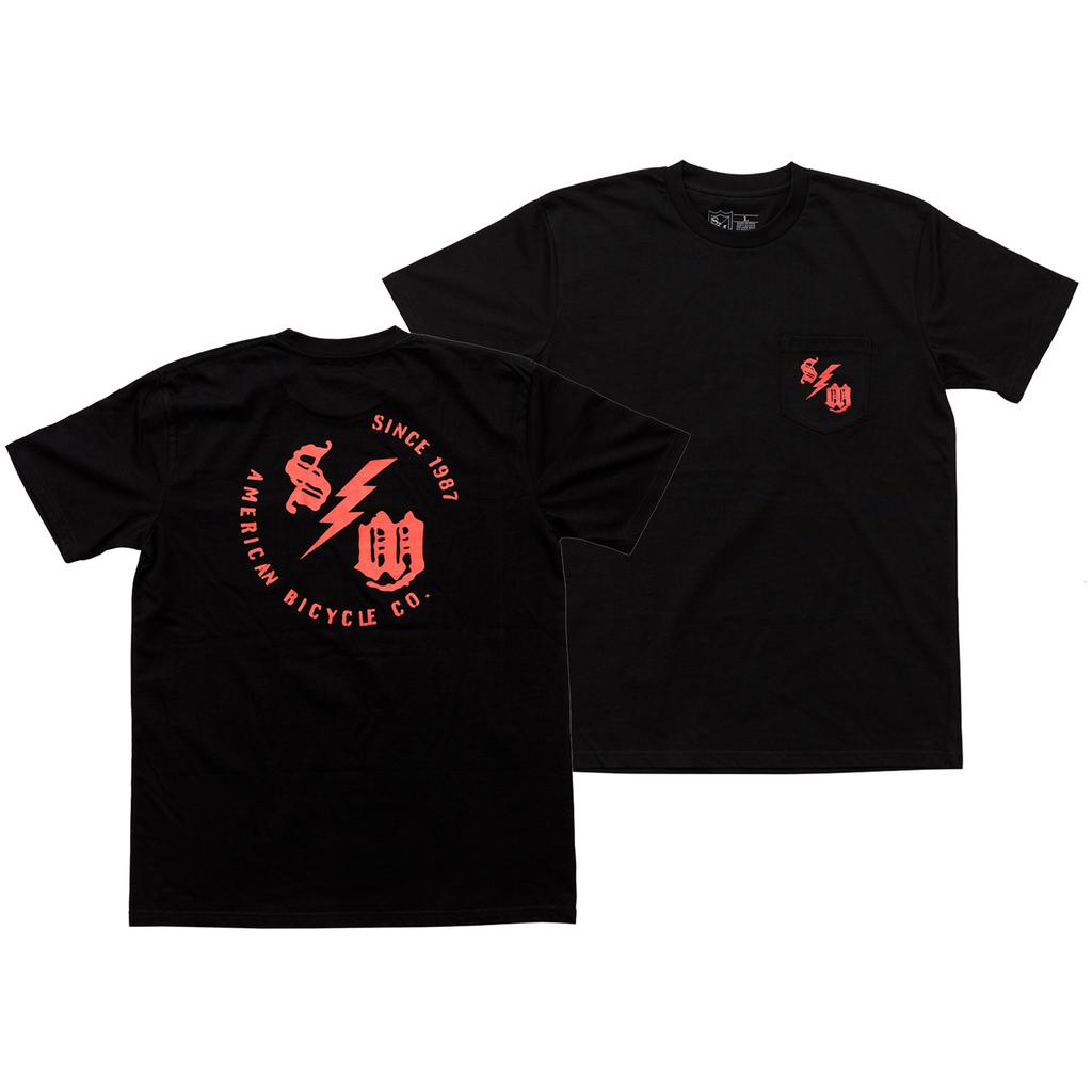 The S&M Bolt Pocket T-Shirt is a black, 100% combed cotton tee with a small red logo on the front pocket and a bold "American Bicycle Co. Since 1981" graphic plus stylized initials on the back, printed in the USA.