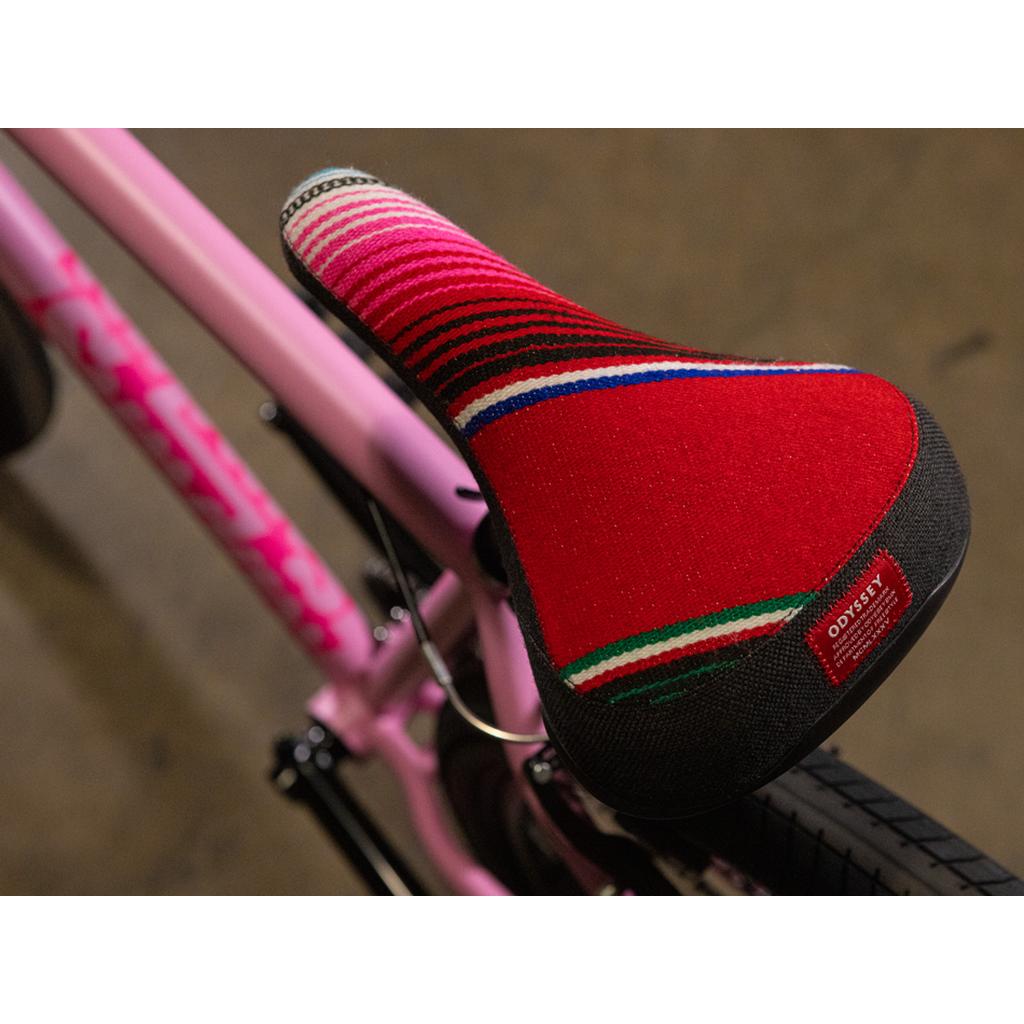 Close-up of the Sunday High C 29 Inch Bike—a pink cruiser bicycle with a vibrant, striped red seat featuring a small Odyssey brand label—set against a blurry brown background. This bike highlights its sturdy chromoly frame, offering both style and durability.
