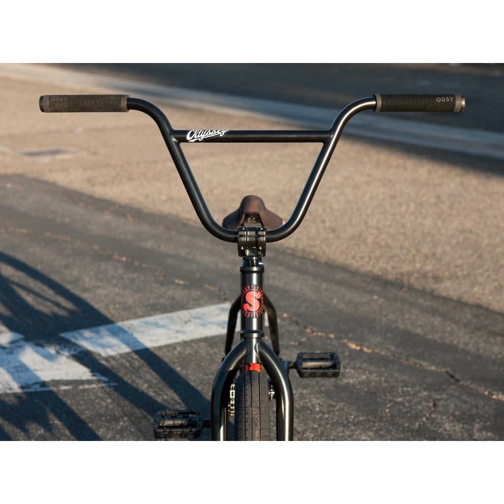 Front view of a black Sunday Sound Wave Special 20 Inch Bike (2023), parked on pavement, showcasing its handlebars, front tire, Odyssey parts, and pedals.