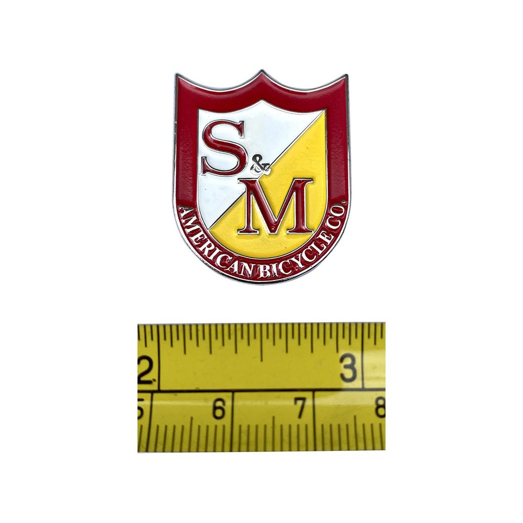 The S&M Shield Pin from S&M American Bicycle Co. boasts a shield logo on an enamel emblem with red, white, and yellow sections and includes a measuring tape for scale. It's a unique addition to any collection.