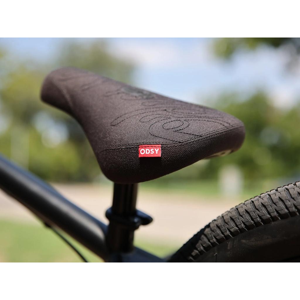 Close-up of a bicycle seat with a red ODYSY tag on the Fairdale Hareraiser FX 26 Inch Bike, showing part of the rear tire and a blurred outdoor background.