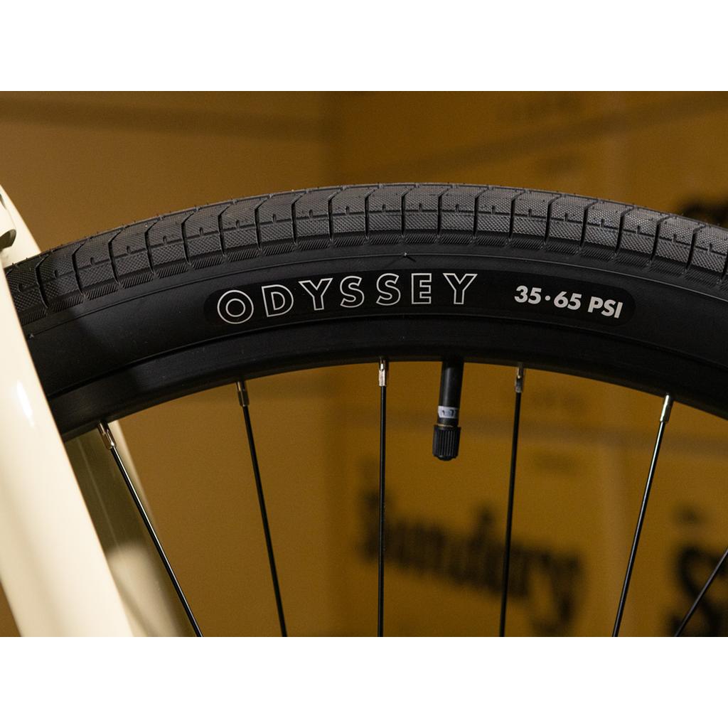 Close-up of a black Odyssey bicycle tire mounted on a wheel, revealing the tread, valve stem, and the 35-65 PSI pressure range printed on the sidewall—ideal for the Sunday High C 29 Inch Bike, which features a strong chromoly frame.