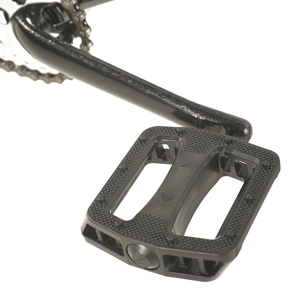 A close-up view of a black bicycle pedal attached to a crank arm on a Division Blitzer 20 Inch Bike. The pedal is rectangular with a textured surface and two open slots. This BMX bike features a high-tensile frame for extra durability.