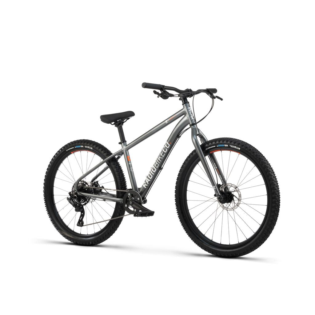 A kid-sized MTB, the Radio 26 Inch Zuma Bike features a lightweight alloy frame in silver with thick tires, a black seat, and front suspension. The handlebars have grips, and the frame is labeled "Raleigh." The bike boasts a gear mechanism and disc brakes for optimal performance.