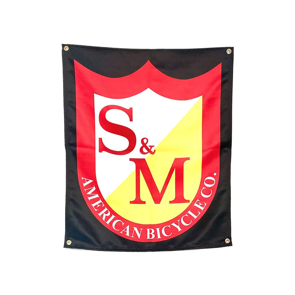 The S&M Shield Banner is a black fabric banner with a red and yellow shield logo, "S&M" and "AMERICAN BICYCLE CO." in white and red text. Proudly made in the USA.