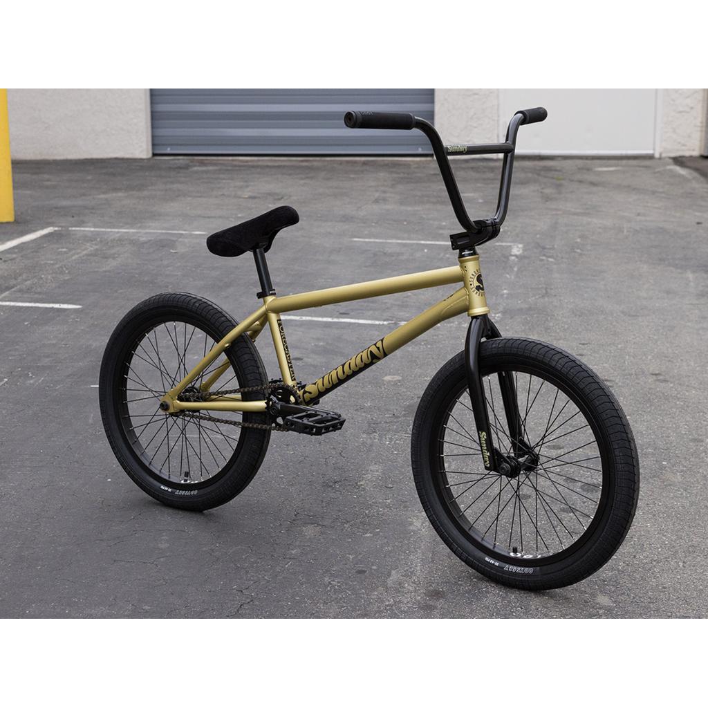 A Sunday Forecaster 20 Inch Bike (2025) / Broc Raiford, featuring a gold 100% chromoly frame with black handlebars, seat, and wheels, is parked on an empty paved lot near a building with a gray garage door.
