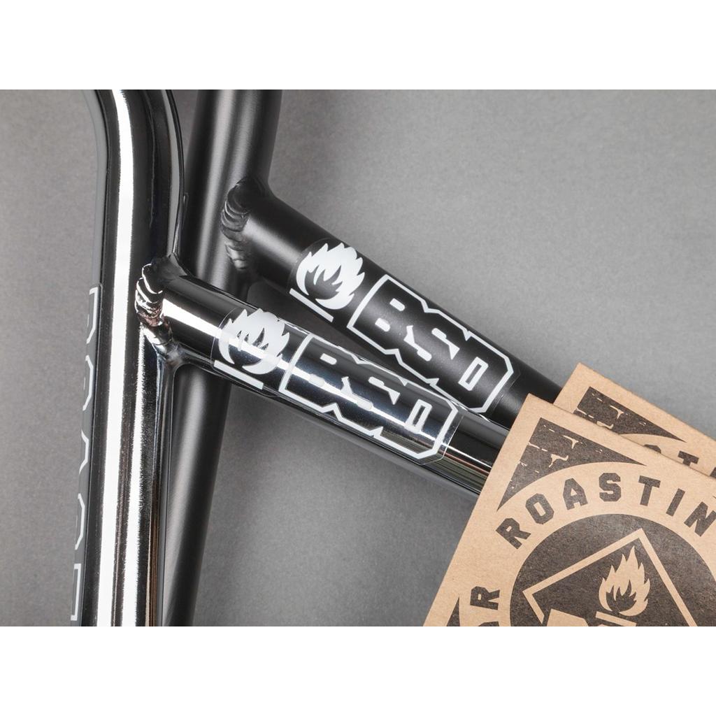 Close-up of a black BMX frame with BSD logo decals, featuring BSD Roastin Bars (Tam Roulston) chromoly handlebars and two brown BSD sticker cards on a gray background.