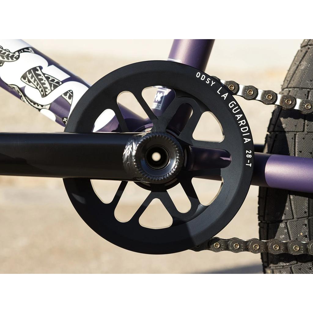 Close-up of a Broc Raiford signature crank and black "ODSY LA GUARDIA 28-T" chainring, showing the chain, rear tire, and part of the 100% Chromoly frame on a Sunday Dark Wave Authentic 20 Inch Bike.