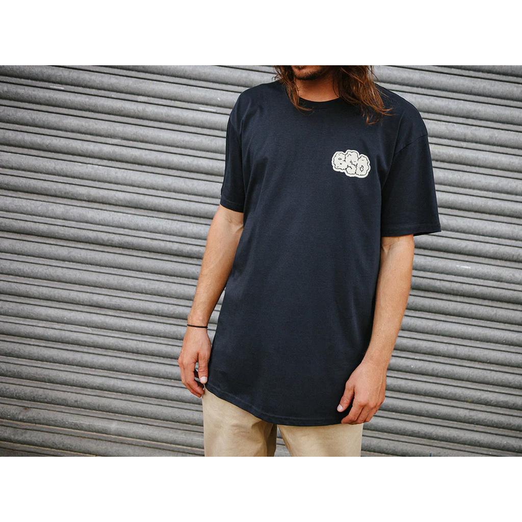 A person in a BSD Burnout T-Shirt, made from lightweight black cotton with a small front logo, stands in front of a corrugated metal wall.