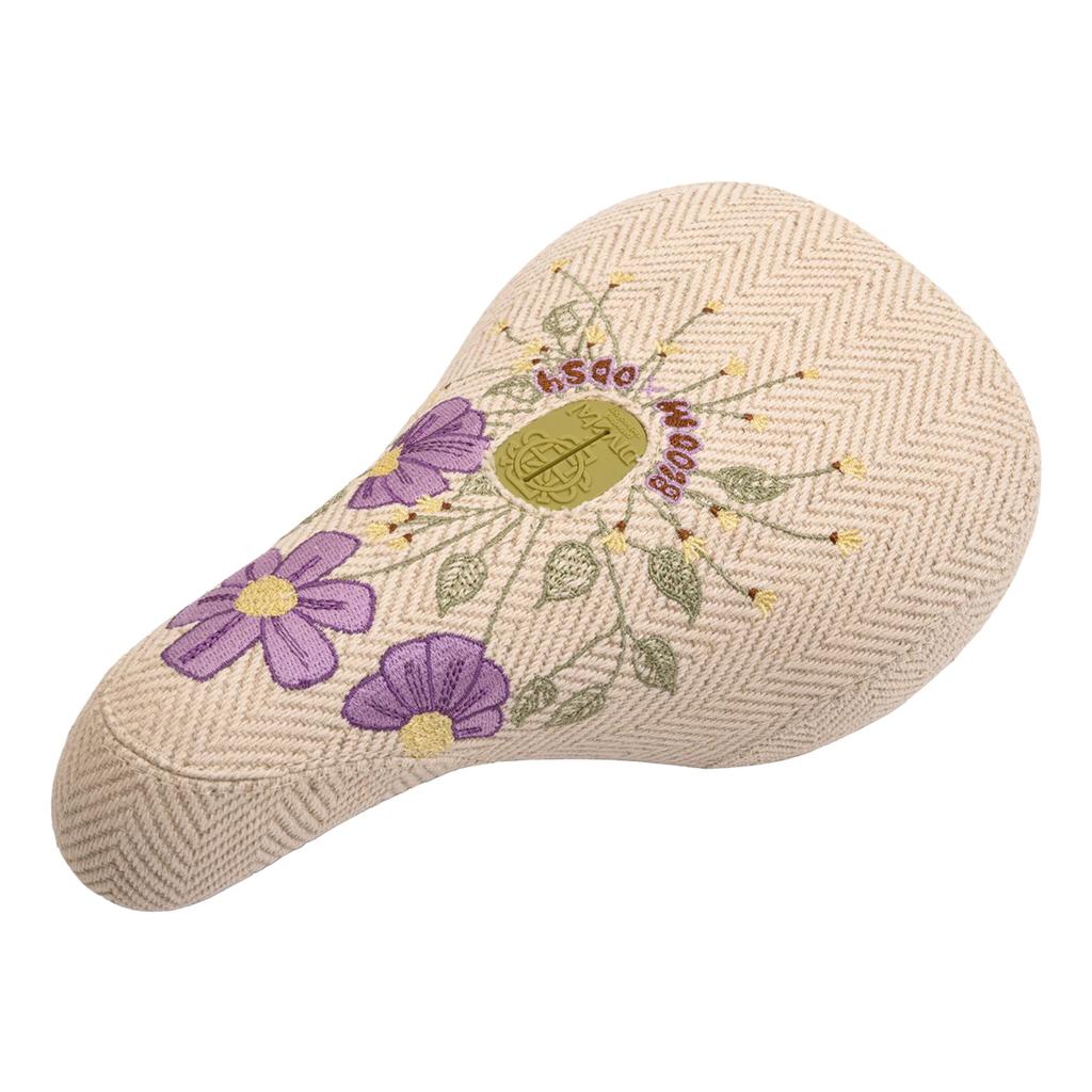 The Odyssey Bloom V2 Pivotal Seat is a beige BMX seat with a herringbone pattern, embroidered purple flowers, green leaves, gold circular emblem, and bold red text, designed for women riders.