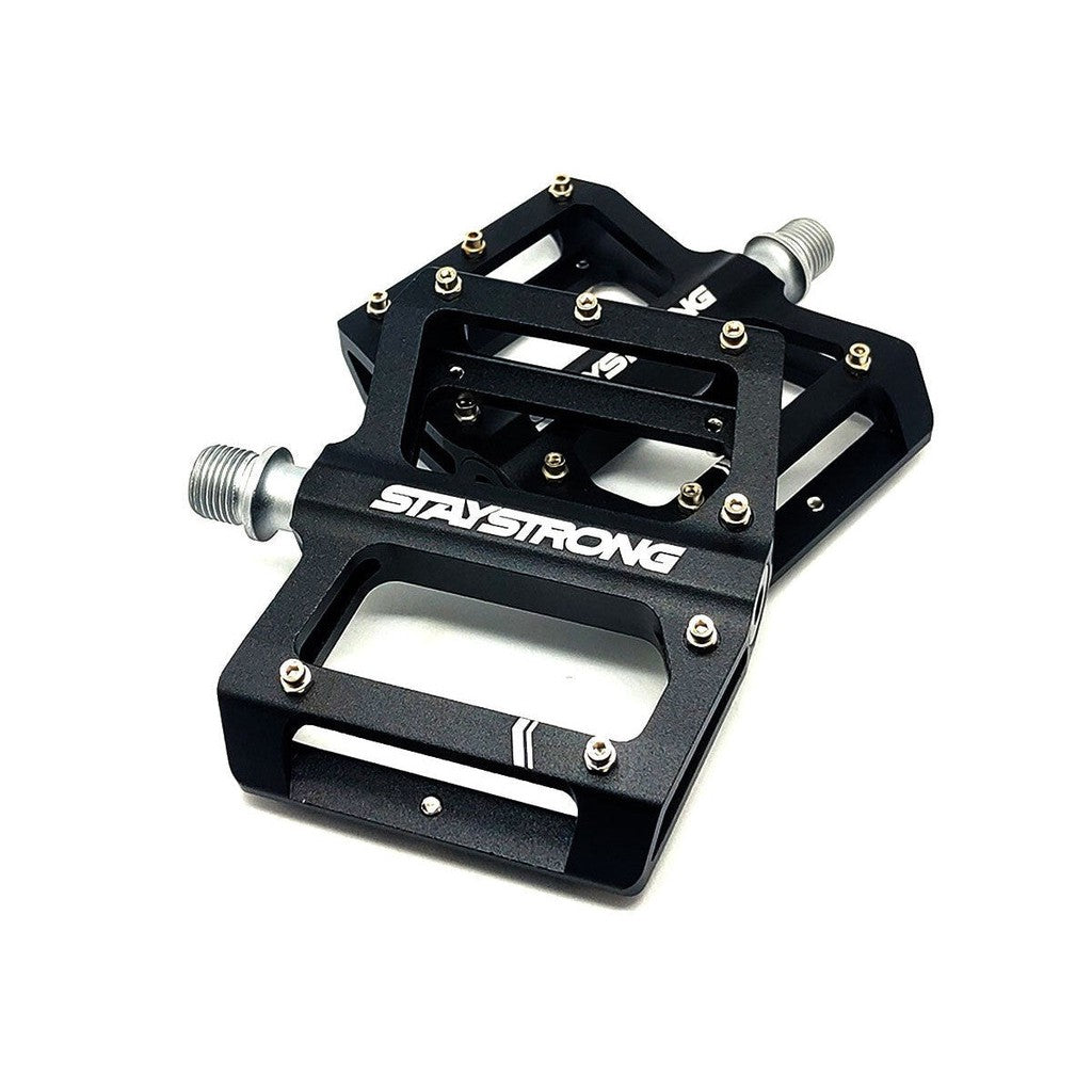 A pair of black Stay Strong PIVOT Junior Platform Pedals with "STAY STRONG" branding, featuring a flat design and multiple small pins for grip, precision crafted using CNC machining.