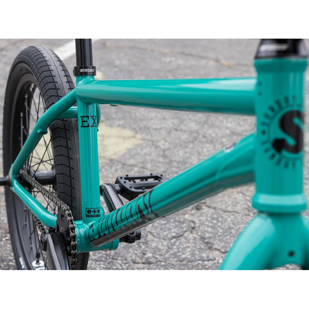 Close-up of the Sunday EX Erik Elstran 20 Inch Bike in turquoise with a durable chromoly frame and black lettering, highlighting the rear wheel, pedals, and seat on an asphalt surface.