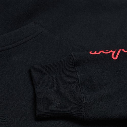 Close-up of the TLD 25.1 Badge Hoodie, a black sweatshirt ideal for winter, showcases red script embroidery on the sleeve. The image highlights the ribbed cuff and fabric texture, merging comfort and style seamlessly.