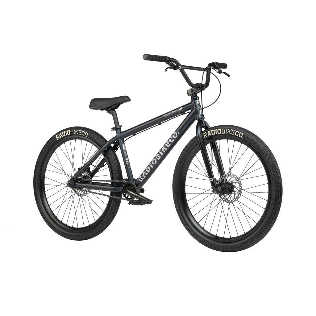 A black Radio 26 Inch Legion Bike with "RADIOBIKECO" written on the tires. The bike features a sleek frame, wide handlebars, knobby tires for off-road use, and Tektro disc brakes for superior stopping power.