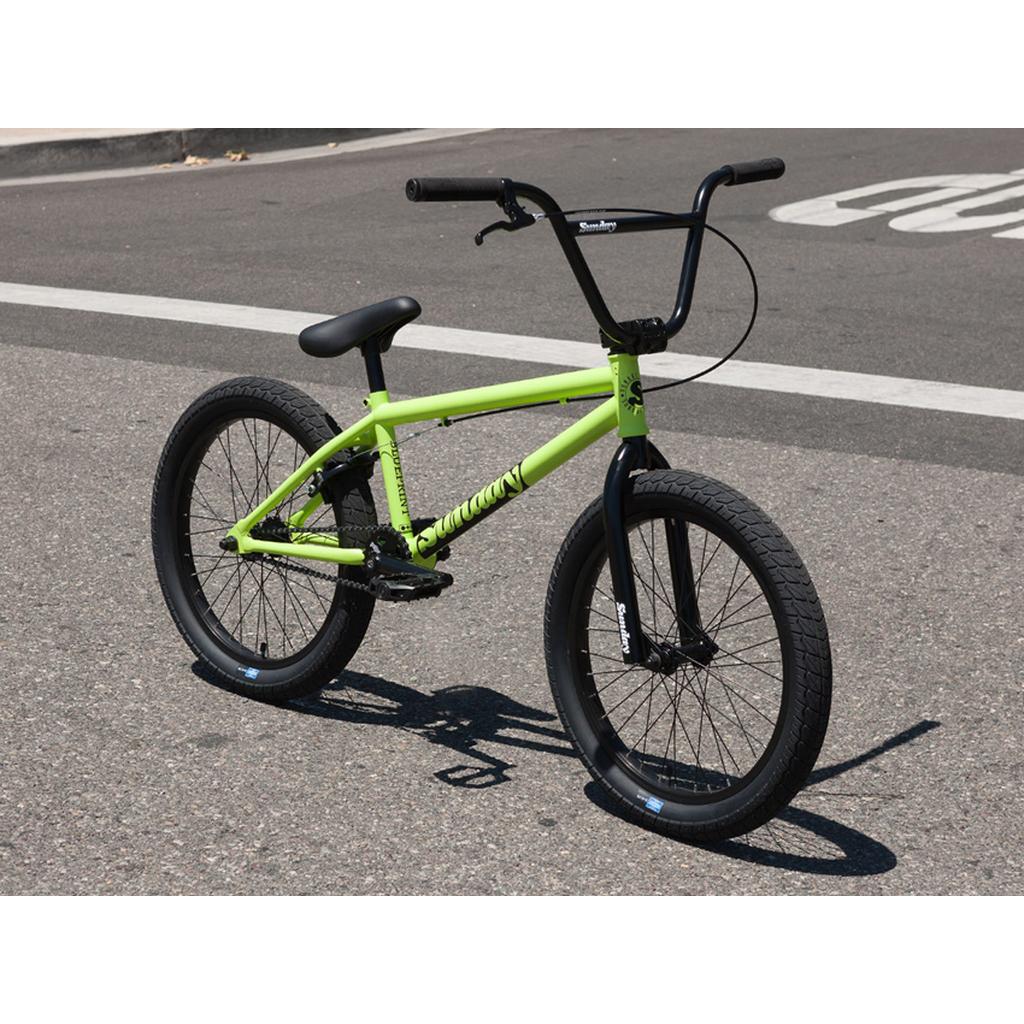 A Sunday Blueprint 20 inch Bike in lime green, equipped with black handlebars and wheels and designed with pro-level geometry perfect for beginner BMX riders, is parked on an asphalt street near a painted crosswalk line.