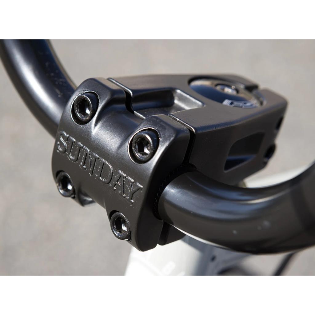 Here’s a rewritten version of your sentence using the provided product data: A close-up view of the black bicycle stem, engraved with the "SUNDAY" brand name and secured with four bolts—an essential part of the Sunday Blueprint 20 inch Bike, which offers beginner-friendly BMX features paired with professional-grade geometry.