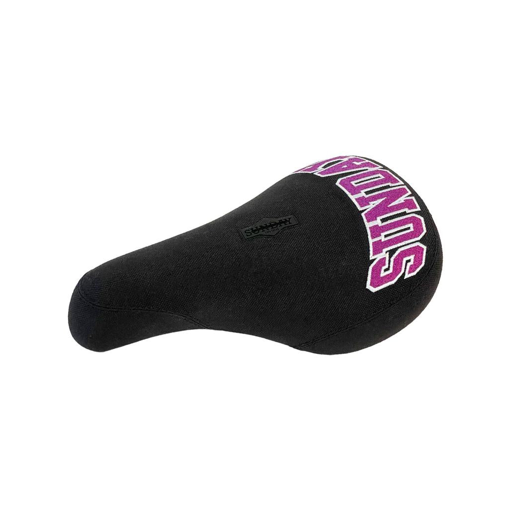 The Sunday Blockhead Pivotal Seat is a black bicycle seat with "SUNDAY" in purple text on top, offering a textured surface and classic saddle shape for ergonomic comfort.