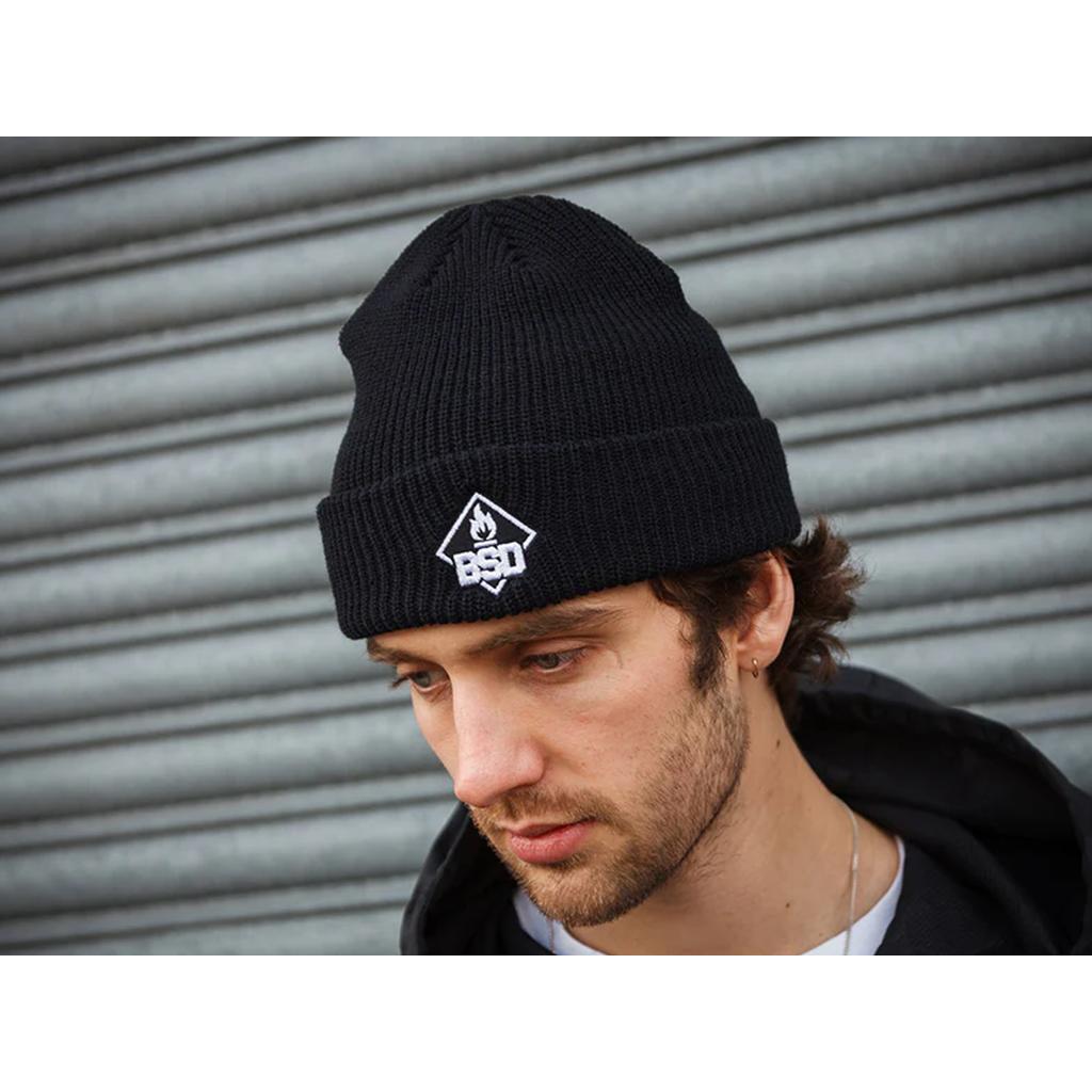 A person is confidently standing in front of a metal shutter, wearing a stylish black BSD Roastin Beanie featuring a bold logo.