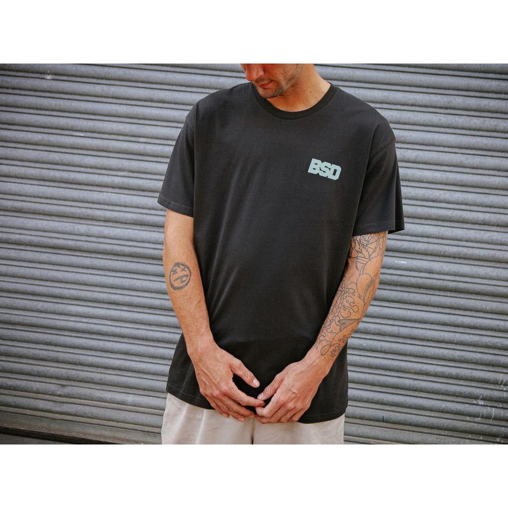 A person wearing a black BSD We Here Forever T-Shirt and light-colored shorts stands in front of a corrugated metal door, with tattoos visible on both arms.