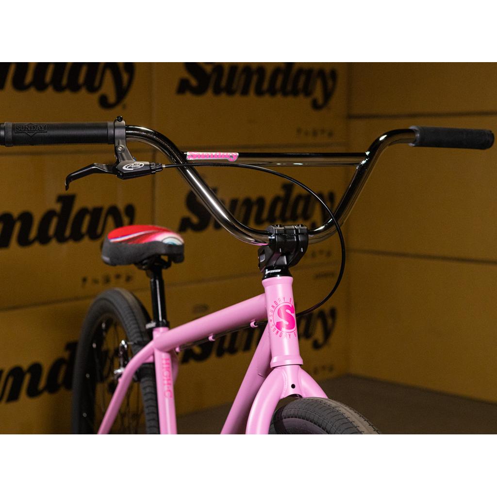 Close-up of the Sunday High C 29 Inch Bike in pink, showcasing its black handlebars and black-and-red seat, set against a backdrop of repeating “Sunday” logos. This stylish cruiser features a robust chromoly frame, delivering both standout looks and exceptional performance.