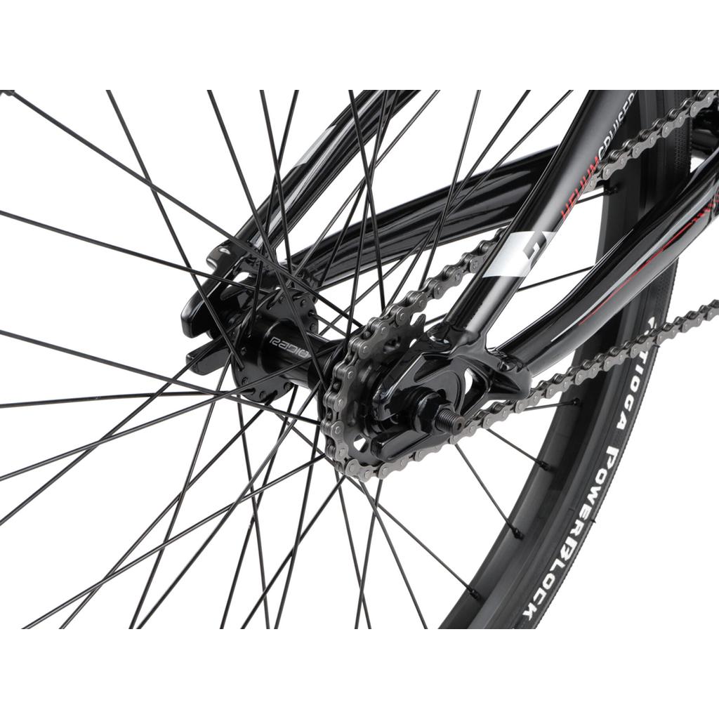 Close-up of the Radio Helium Pro 24 Inch Cruiser Bike’s rear wheel, highlighting the chain, gears, spokes, hub, and a section of the tire marked "TIOGA POWERBLOCK.