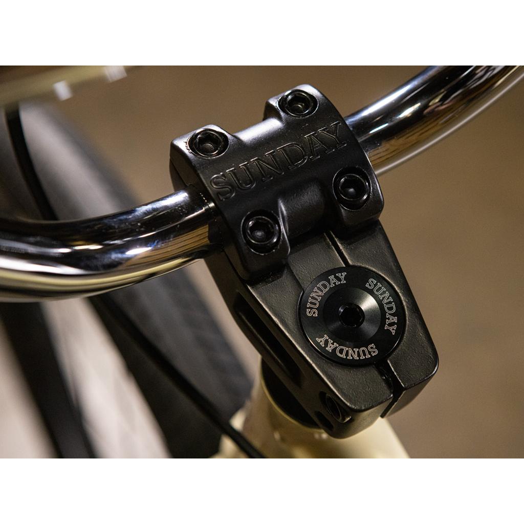 Close-up of a black stem clamp and handlebars engraved with "SUNDAY," highlighting bolt details and part of the front tire on the Sunday High C 29 Inch Bike—a sleek 29" cruiser engineered for strength with its rugged chromoly frame.
