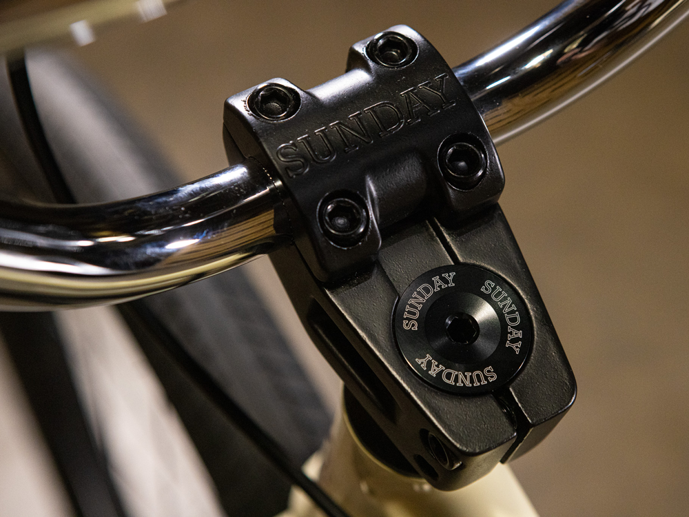 Close-up of a black stem clamp and handlebars engraved with "SUNDAY," highlighting bolt details and part of the front tire on the Sunday High C 29 Inch Bike—a sleek 29" cruiser engineered for strength with its rugged chromoly frame.