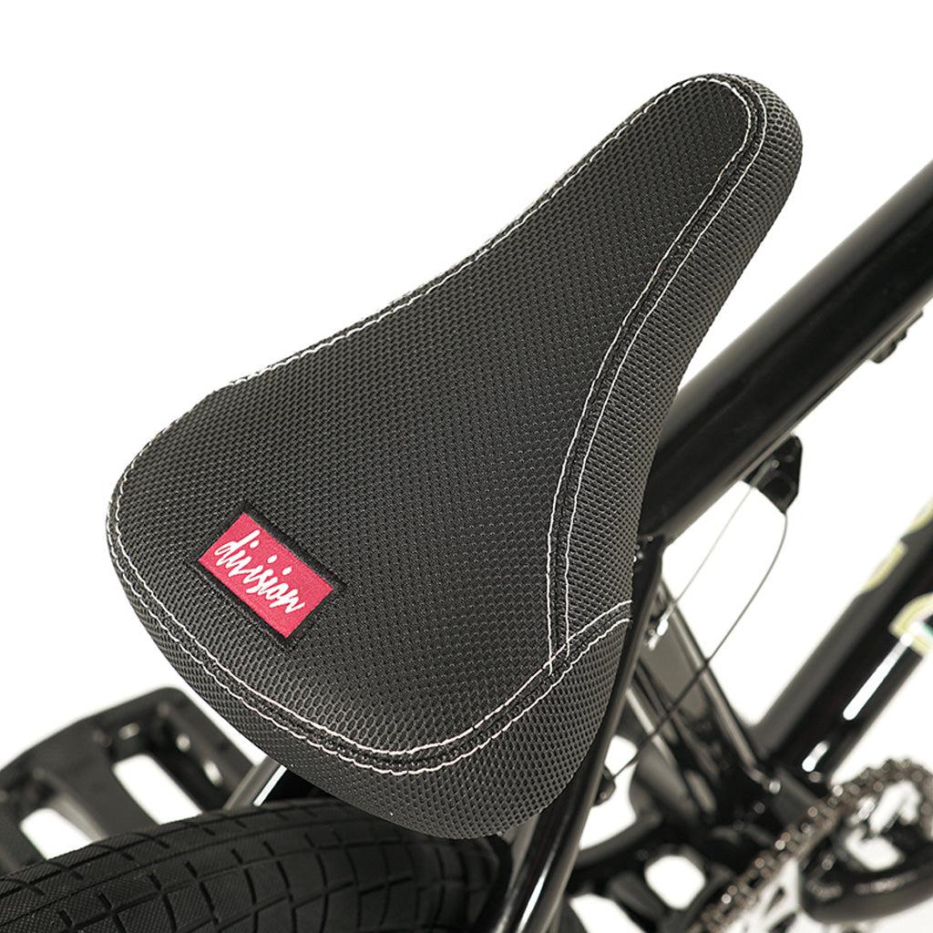 Close-up image of the black bicycle seat with white stitching and a textured surface, featuring a red and white label. The back wheel and part of the hi-tensile frame of the Division Blitzer 20 Inch Bike are visible.
