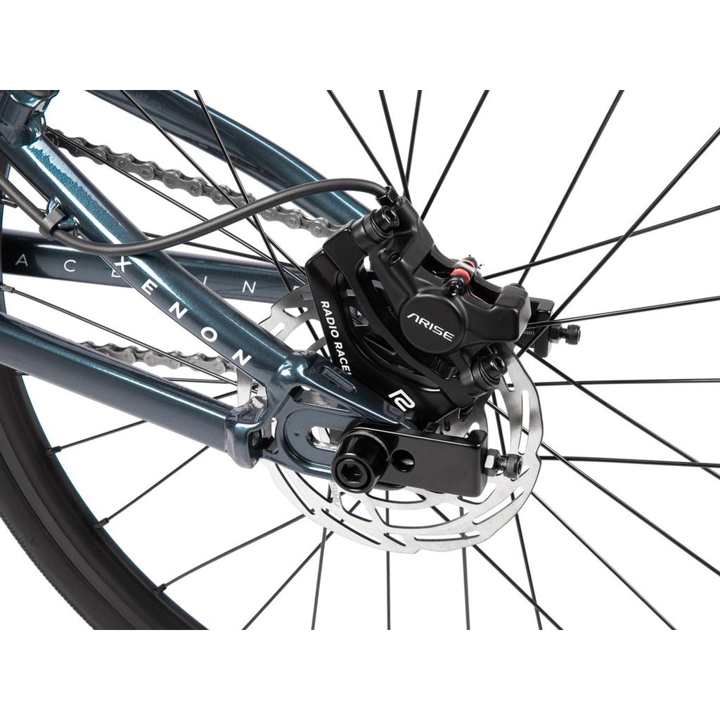 Close-up of the rear wheel of the Radio Xenon Junior Disc 20 Inch Bike, showcasing its disc brake system with visible rotor and caliper, as well as a portion of its hydroformed 6061-T6 alloy frame labeled "XENON." The image also highlights the sturdy Tioga Power Block tyres.