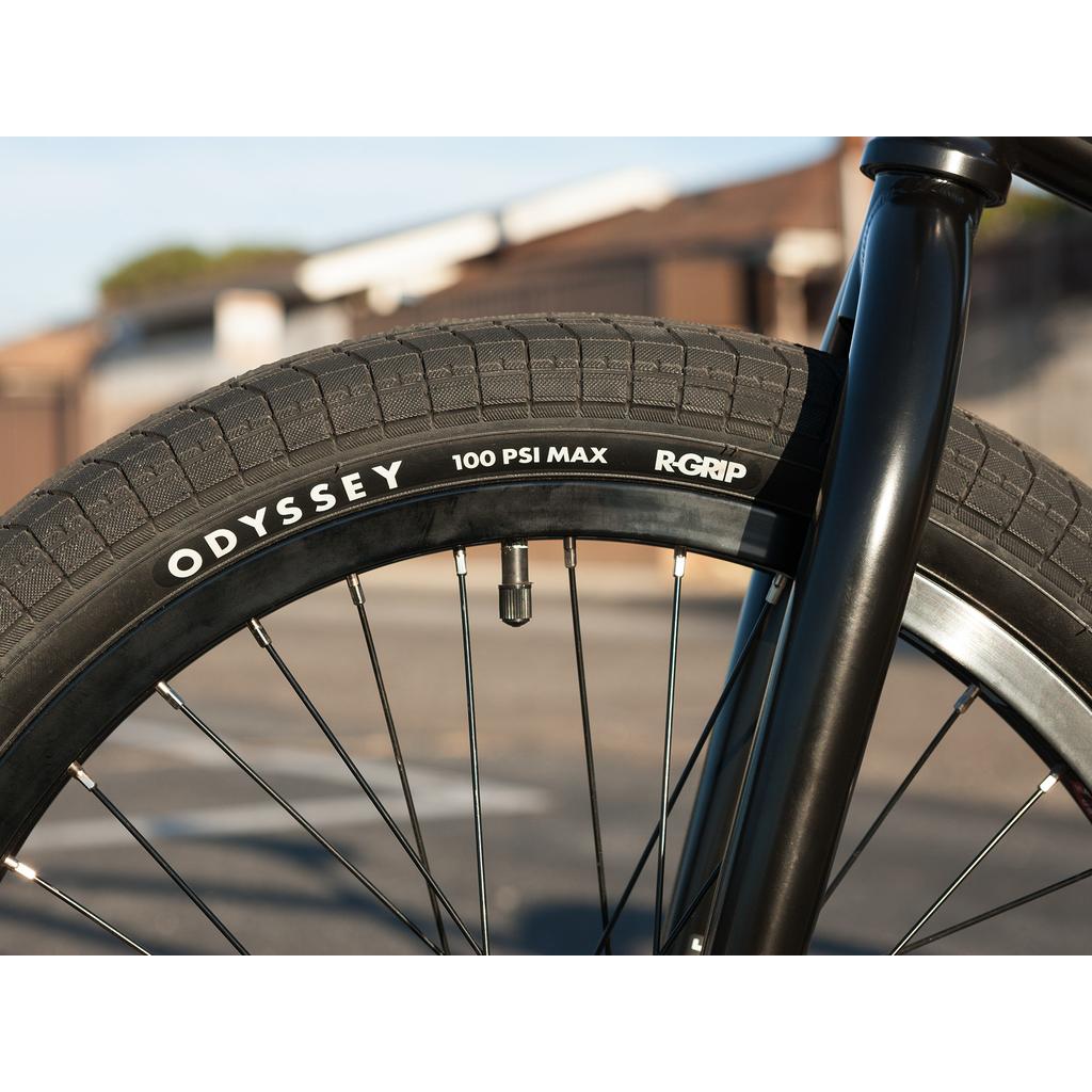 Close-up of a Sunday Sound Wave Special 20 Inch Bike (2023) front wheel with an Odyssey tire featuring "100 PSI MAX" and "R-GRIP" markings, parked outdoors on a sunny day—ideal for top-tier BMX riding.