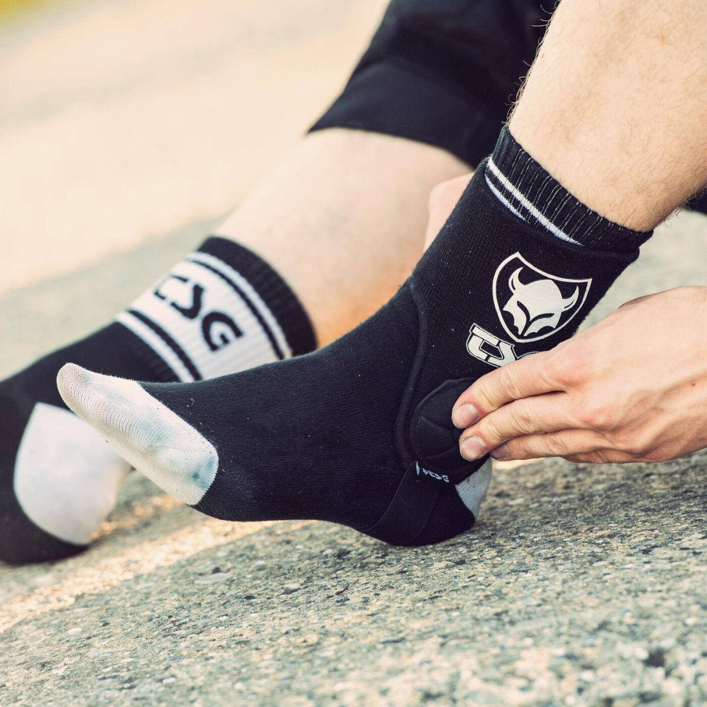 A person adjusts a black TSG Ankle Guard Cam sock with a white logo and EVA foam ankle pads for extra protection; the other foot wears the matching sock with gray accents.
