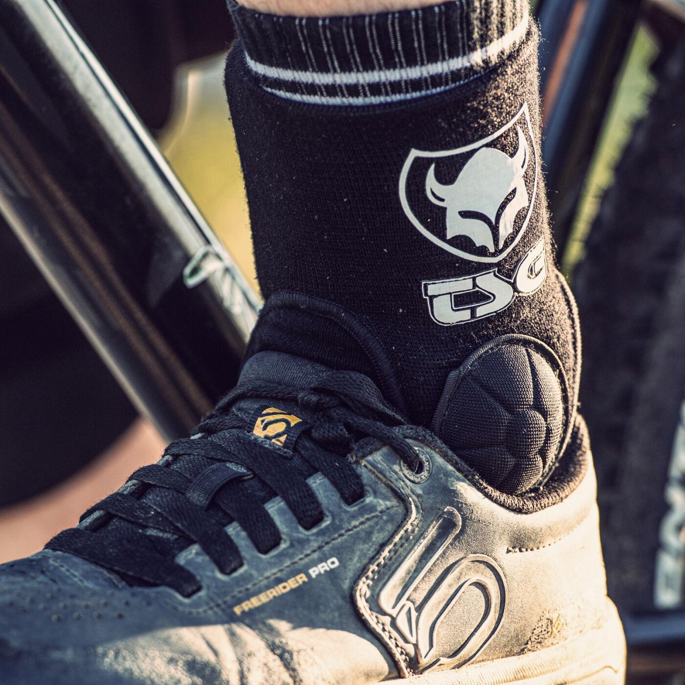 Close-up of a person wearing black socks with a white logo and the TSG Ankle Guard Cam, showcasing EVA foam ankle pads for added protection, next to a bicycle tire.