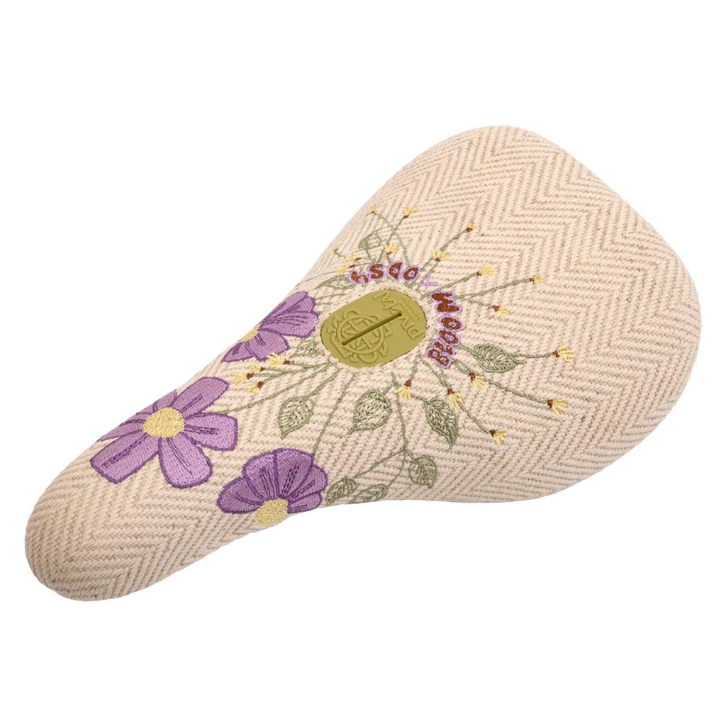 The Odyssey Bloom V2 Pivotal Seat features a cream color, herringbone pattern, purple embroidered flowers, and a gold circular logo at the center—crafted to inspire women riders.