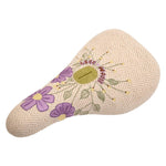 The Odyssey Bloom V2 Pivotal Seat features a cream color, herringbone pattern, purple embroidered flowers, and a gold circular logo at the center—crafted to inspire women riders.