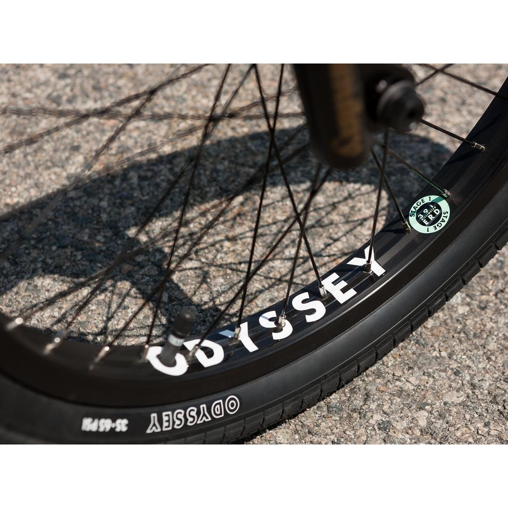 Close-up of a bicycle wheel with black spokes and an "ODYSSEY" tire on pavement, part of the Sunday Forecaster 20 Inch Bike (2025) / Broc Raiford, featuring an Odyssey Clutch v2 Freecoaster.
