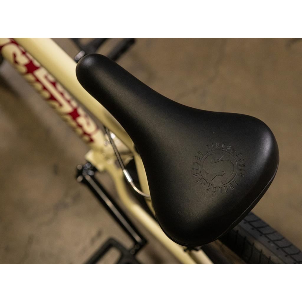 Close-up of a black seat featuring embossed branding, mounted on the cream-colored chromoly frame of the Sunday High C 29 Inch Bike, resting on a concrete floor.