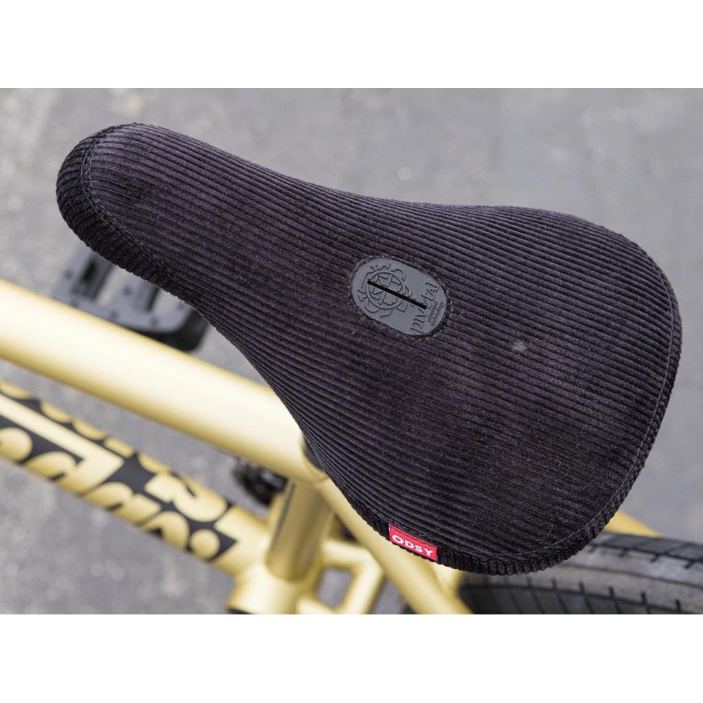 Close-up of a black corduroy BMX seat with an Odyssey logo on the Sunday Forecaster 20 Inch Bike (2025) / Broc Raiford, mounted to a gold 100% chromoly frame, photographed from above.