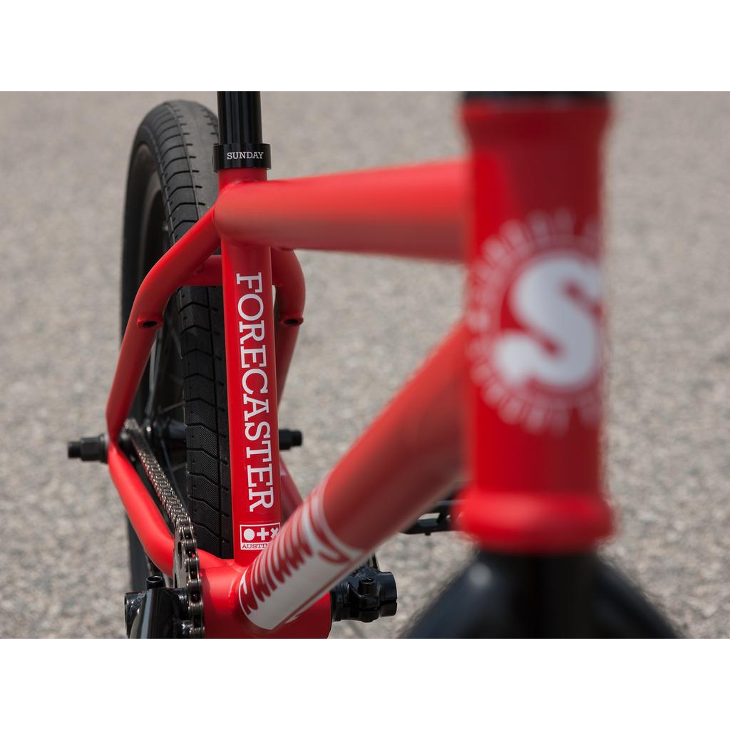 Close-up of the Sunday Forecaster 20 Inch Bike (2025) / Brett Silva in red, showing "Forecaster" and "Sunday" on its 100% chromoly frame and seat tube, set against a blurred paved background.