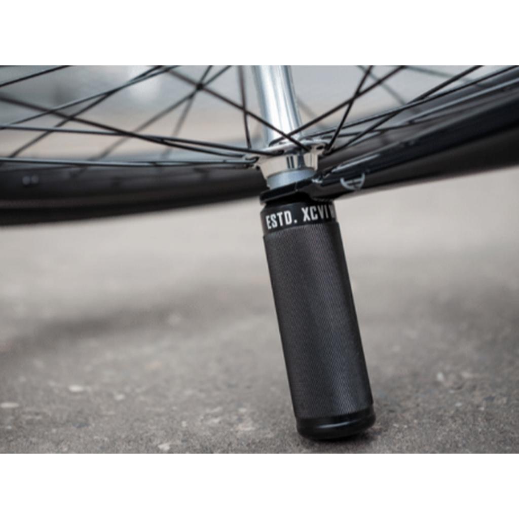 Close-up of a bicycle wheel with metal spokes and a pair of Wethepeople Sinus Flatland Pegs attached to the axle, resting on a concrete surface.
