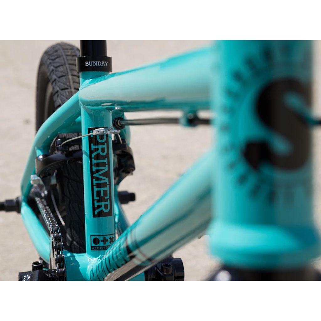 Close-up view of the turquoise frame on the Sunday Primer 16 Inch Bike (2025), highlighting the branding "Primer" and "Sunday," with a focus on the chain and Odyssey Springfield Brakes.
