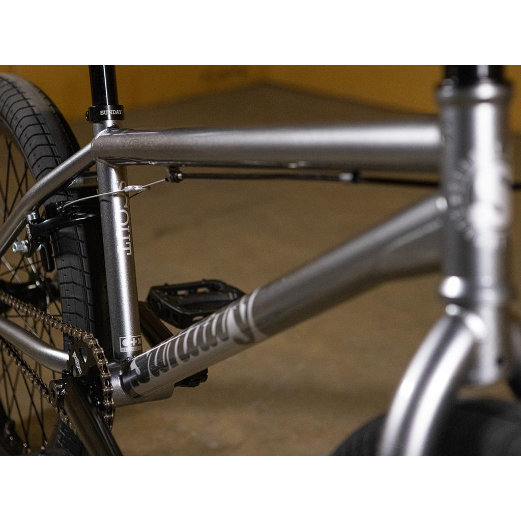 Close-up view of the Sunday Scout 20 Inch Bike’s silver frame, featuring Odyssey parts on the chain, pedals, rear wheel, and seat post against a blurred indoor background.