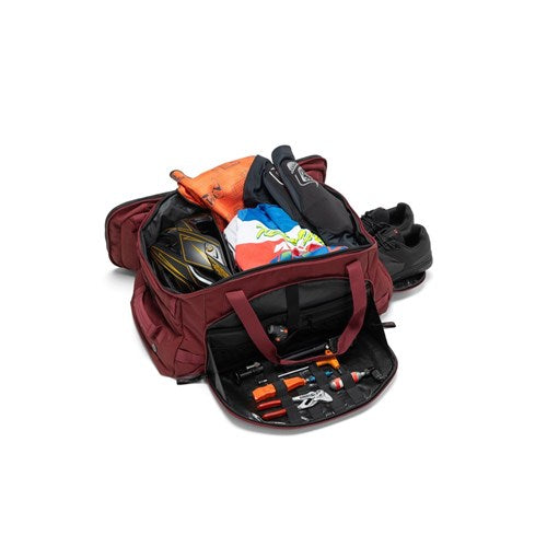 The Albek Skytrail 51 Gear Bag, shown in maroon, features multiple compartments and is displayed open with clothes, shoes, a helmet, tools, and other essentials for organized gear storage.