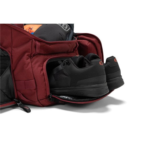 A maroon Albek Skytrail 51 Gear Bag with an open side compartment reveals a pair of black sneakers neatly stored inside, perfect for convenient gear organization.