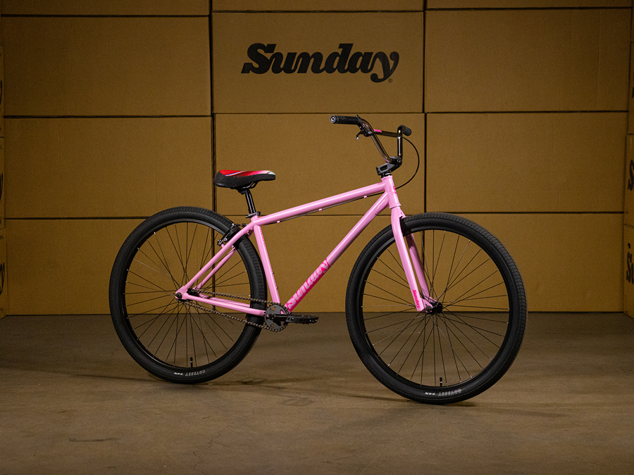 A pink Sunday High C 29 Inch Bike with black tires and a seat featuring red accents is showcased indoors in front of a wall of "Sunday" branded cardboard boxes. This striking display highlights the Sunday High C 29 Inch Bike, recognized for its robust chromoly cruiser frame and high-quality Odyssey components.