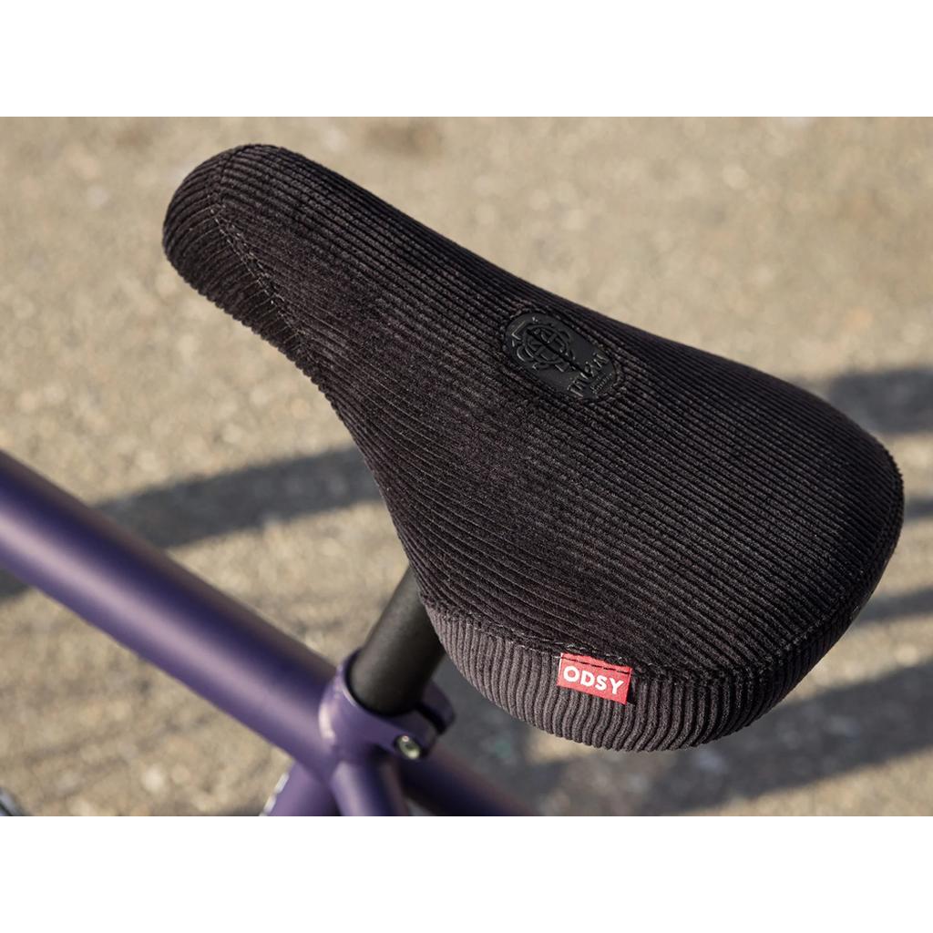 A close-up of a black textured bicycle seat with a red "ODSY" tag, mounted on a purple Sunday Dark Wave Authentic 20 Inch Bike frame.