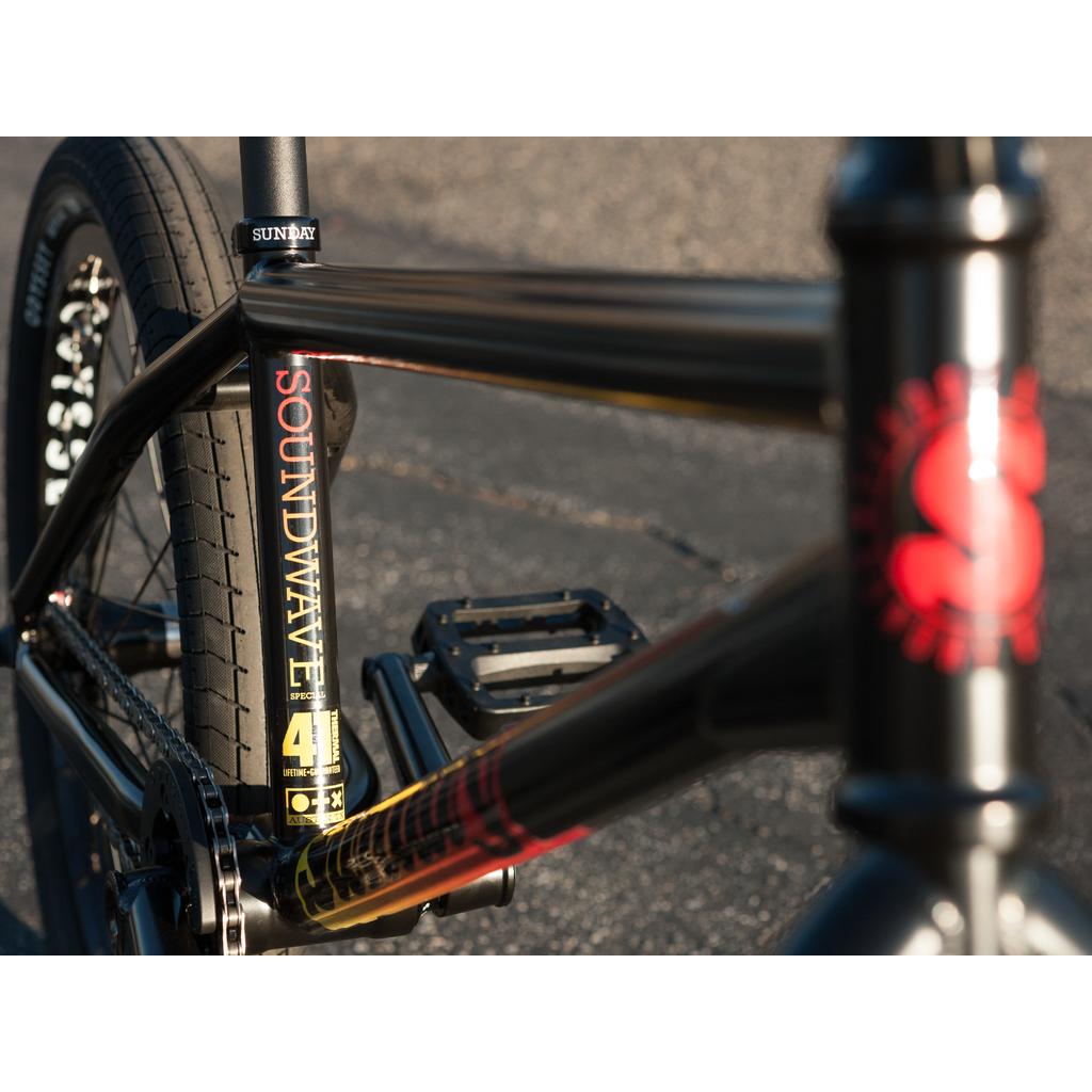 Close-up of a black Sunday Sound Wave Special 20 Inch Bike (2023) BMX, showing the "SOUNDWAVE" frame label, rear wheel, pedal, Odyssey components, part of the headset with a red logo, and pavement background.