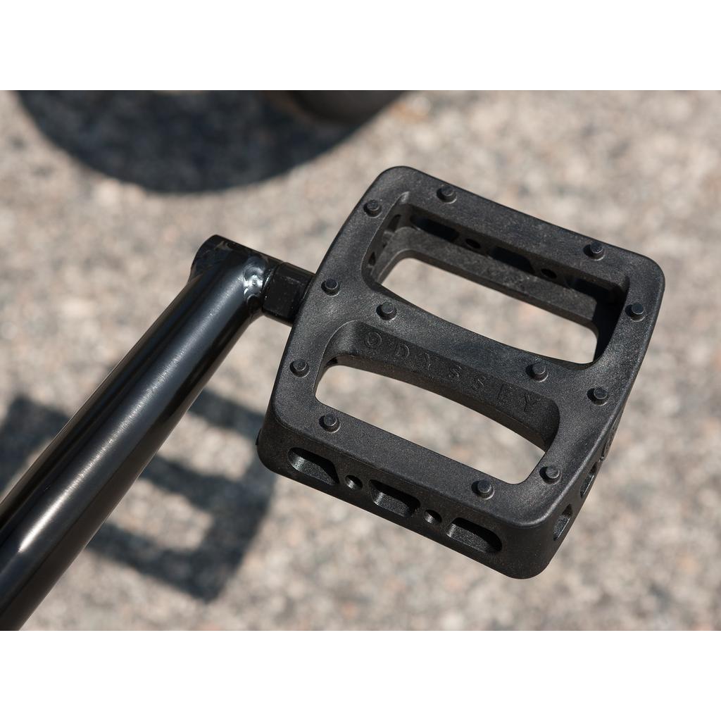 Close-up of a black metal pedal attached to a crank arm on the Sunday Forecaster 20 Inch Bike (2025) / Broc Raiford, featuring a textured surface and visible branding. Gravel ground is in the background.