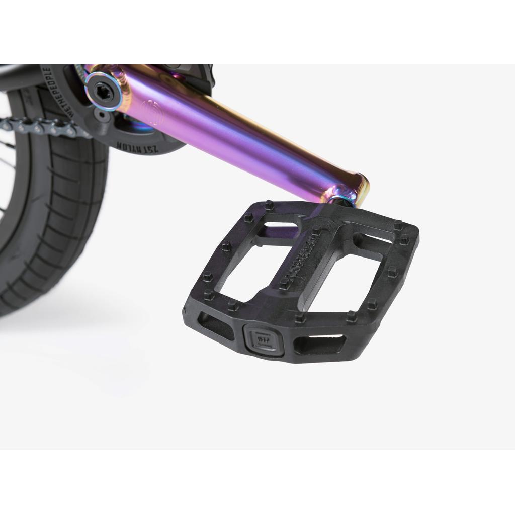 Close-up of a black pedal attached to a rainbow-colored crank arm on the Wethepeople Reason Shift 20 Inch Bike, with part of the chain and tire in the background.