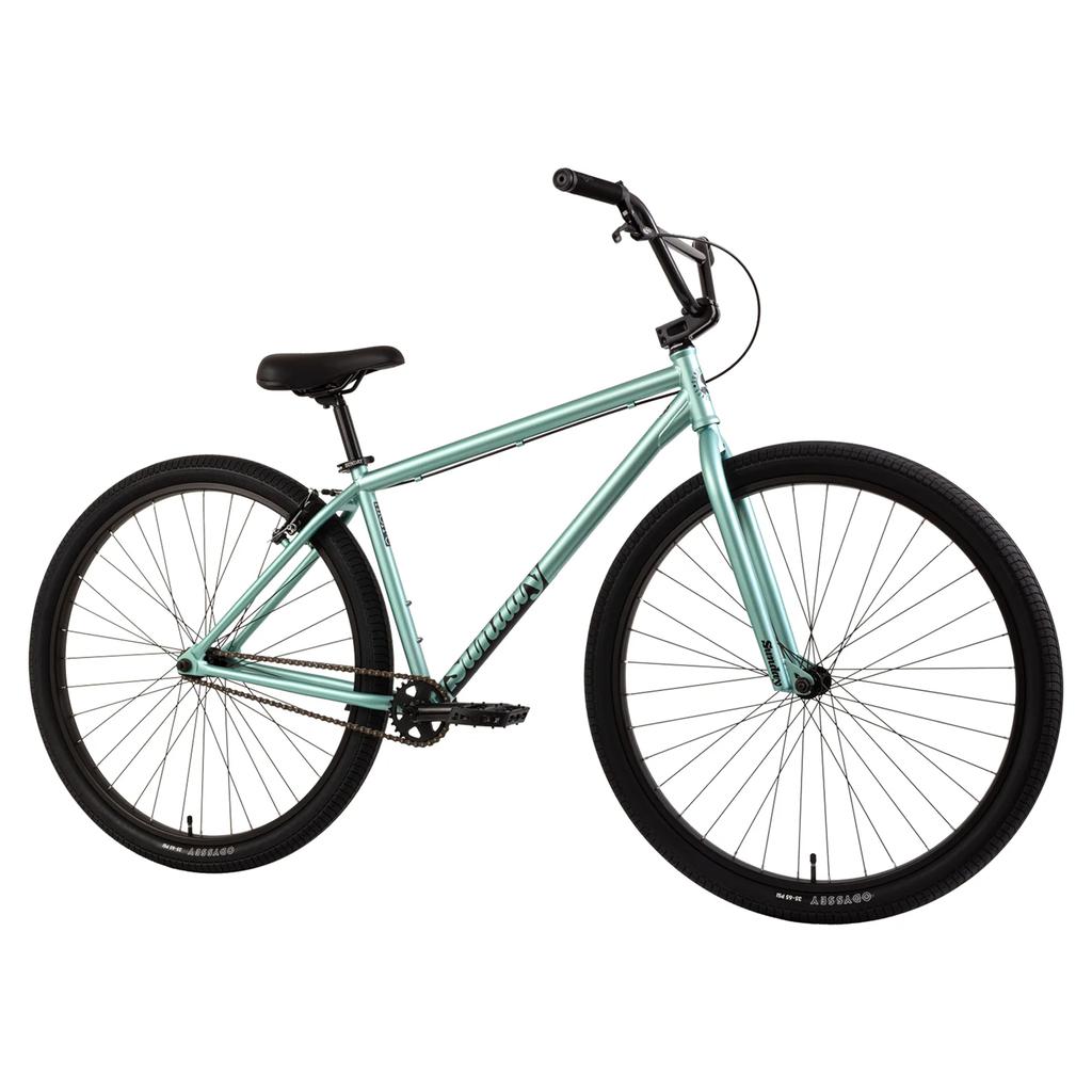 The Sunday High C 29 Inch Bike (2025) is a light blue single-speed bicycle with black handlebars, saddle, and tires, featuring classic cruiser geometry, shown from the side on a white background.