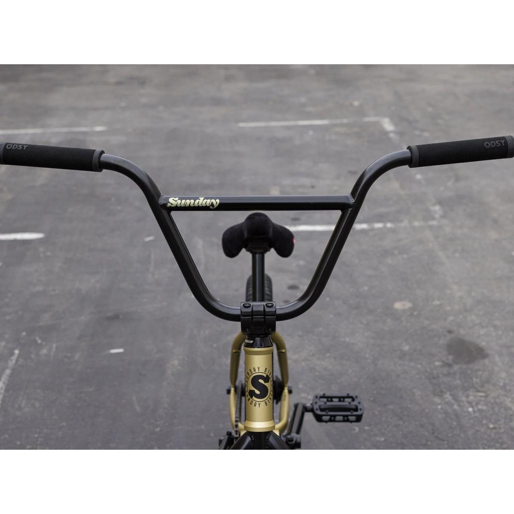 Close-up of the Sunday Forecaster 20 Inch Bike (2025) / Broc Raiford handlebars and front frame, showing "Sunday" and "ODSY" logos on a 100% chromoly frame against an empty paved surface.